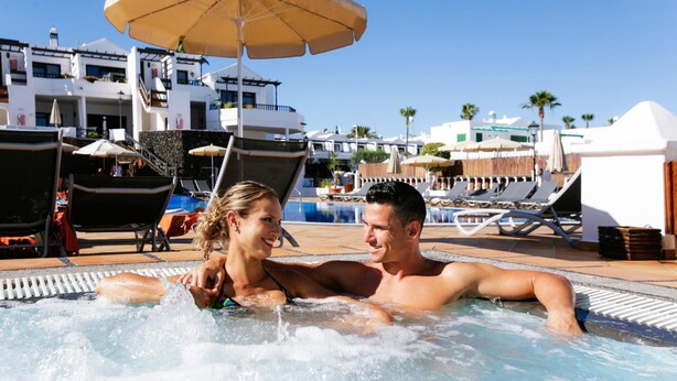 A couple relaxing in a hot tub with a poolside view, lounge chairs, umbrellas, and a building with balconies in the background. - Princesa Ico (Tías)