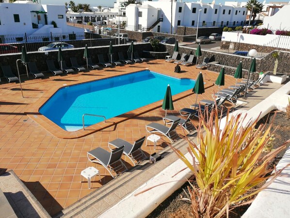 A rectangular swimming pool with a tiled deck, surrounded by lounge chairs and umbrellas, set against a backdrop of white buildings and a clear sky.