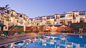 A swimming pool area with lounge chairs and umbrellas, surrounded by buildings with multiple floors and balconies. - Princesa Ico (Tías)