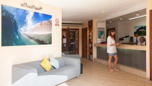 A reception area with a receptionist, a grey sofa with cushions, a large wave painting, and a 'No Smoking' sign. - Princesa Ico (Tías)