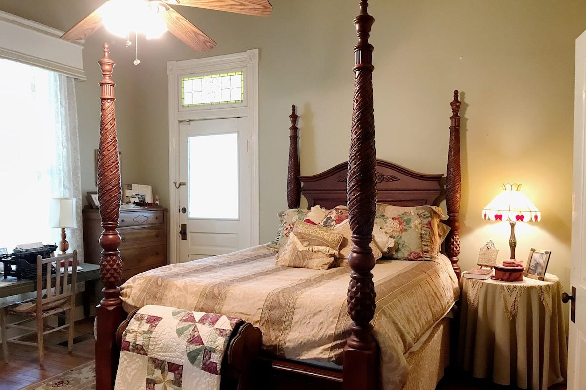 A four-poster bed with a floral bedspread, a nightstand with a lamp, and a view of the outside door.