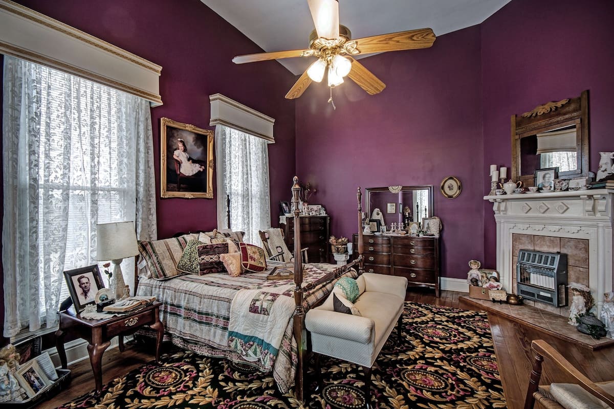 A bedroom with a four-poster bed, a sitting area with a chair, a fireplace, and a ceiling fan.