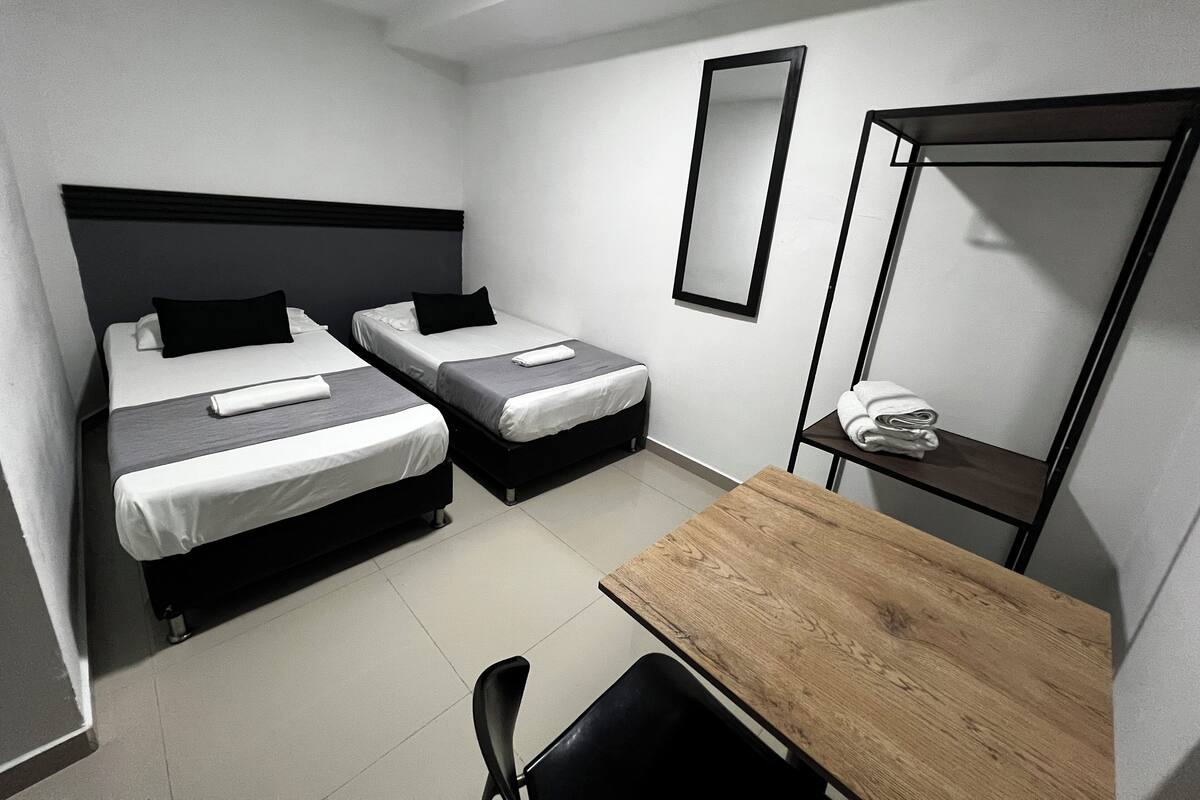 A small, modern hotel room with two single beds, a wooden table, a chair, a mirror, and a shelf with towels.