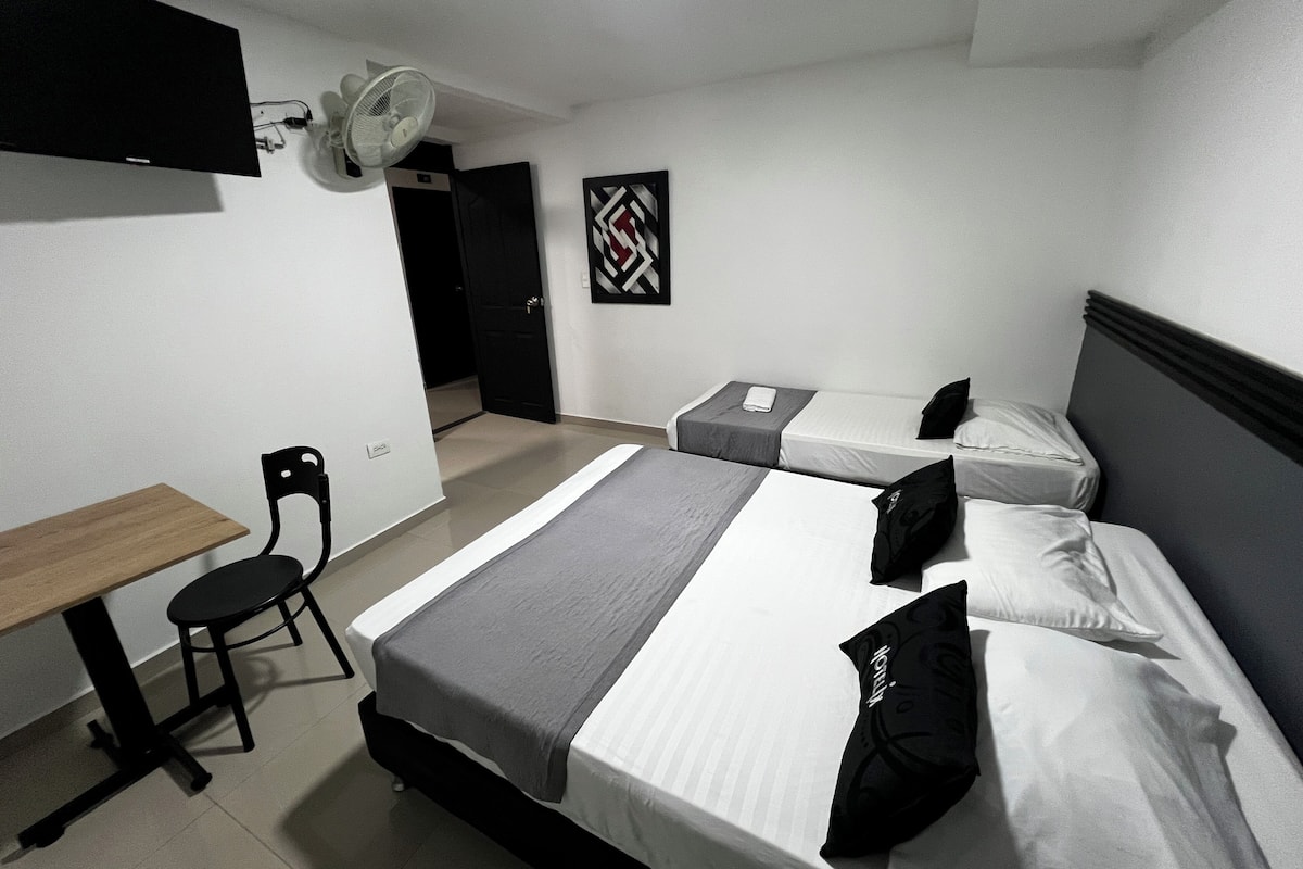 A hotel room with two beds, a desk, a chair, a TV, and a ceiling fan.