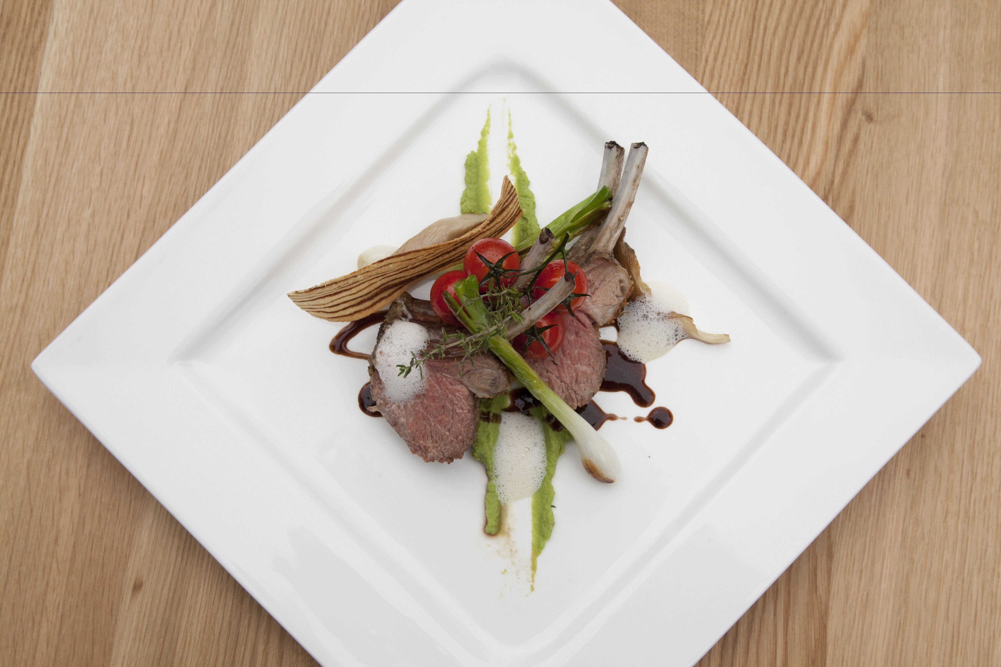 A square white plate with a dish of meat, vegetables, and sauce on a wooden table.