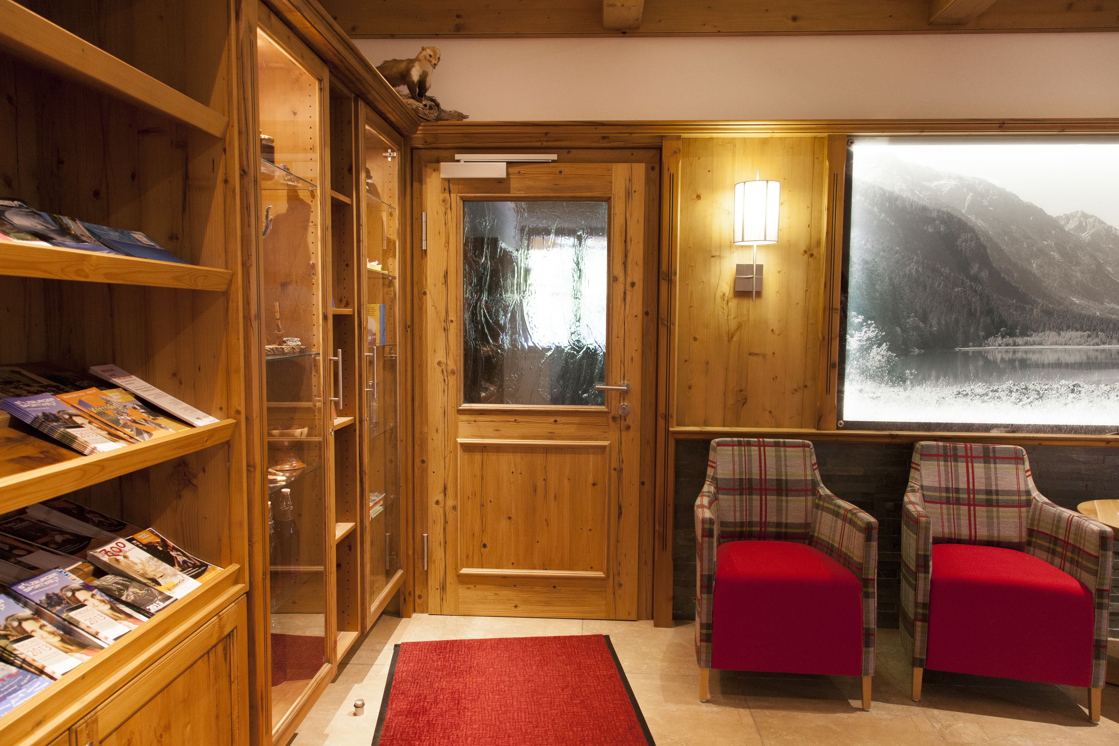 A wooden interior with a fireplace, a bookshelf, two plaid chairs, and a large window with a mountain view.