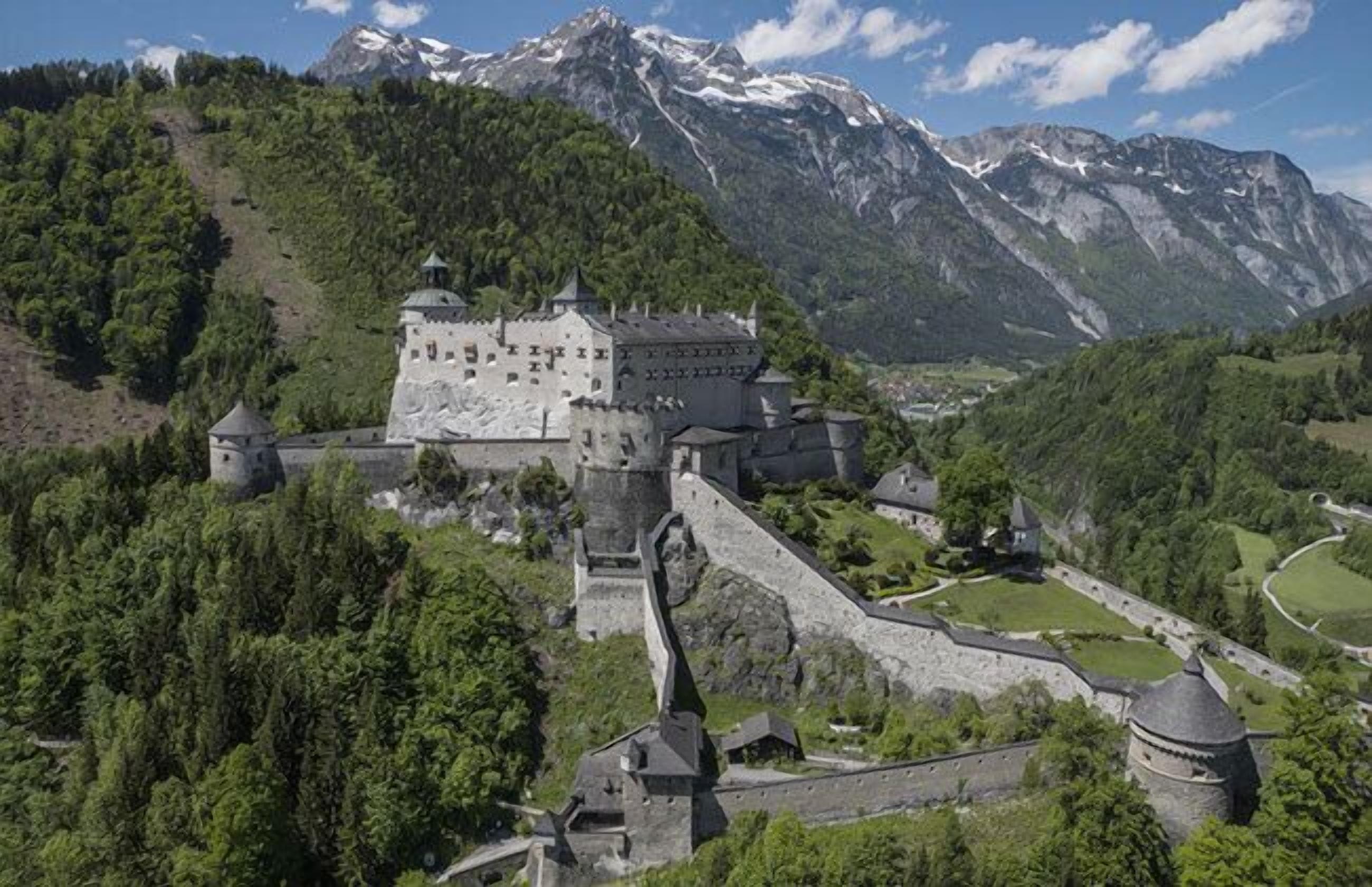 A historic castle with a fortified wall, surrounded by lush greenery and mountains in the background.