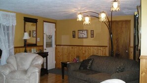 A living room with a beige armchair, a grey sofa, and a white ottoman. There is a wooden floor, a lamp, and a door with a white curtain. - Gullivers Cove Oceanview Cottages (Gullivers Cove)