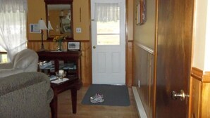 A room with a wooden door, a small table with a lamp, a chair, and a rug leading to the door. - Gullivers Cove Oceanview Cottages (Gullivers Cove)