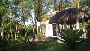 Un jardín tropical con una estructura de techo de paja, palmeras y un edificio blanco con balcón.