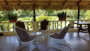 A covered outdoor seating area with wicker chairs and a table, surrounded by potted plants and a view of lush greenery.