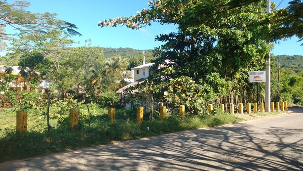 A rural road with a sign for Hotel Casa del SNL, surrounded by lush greenery and a thatched-roof building.