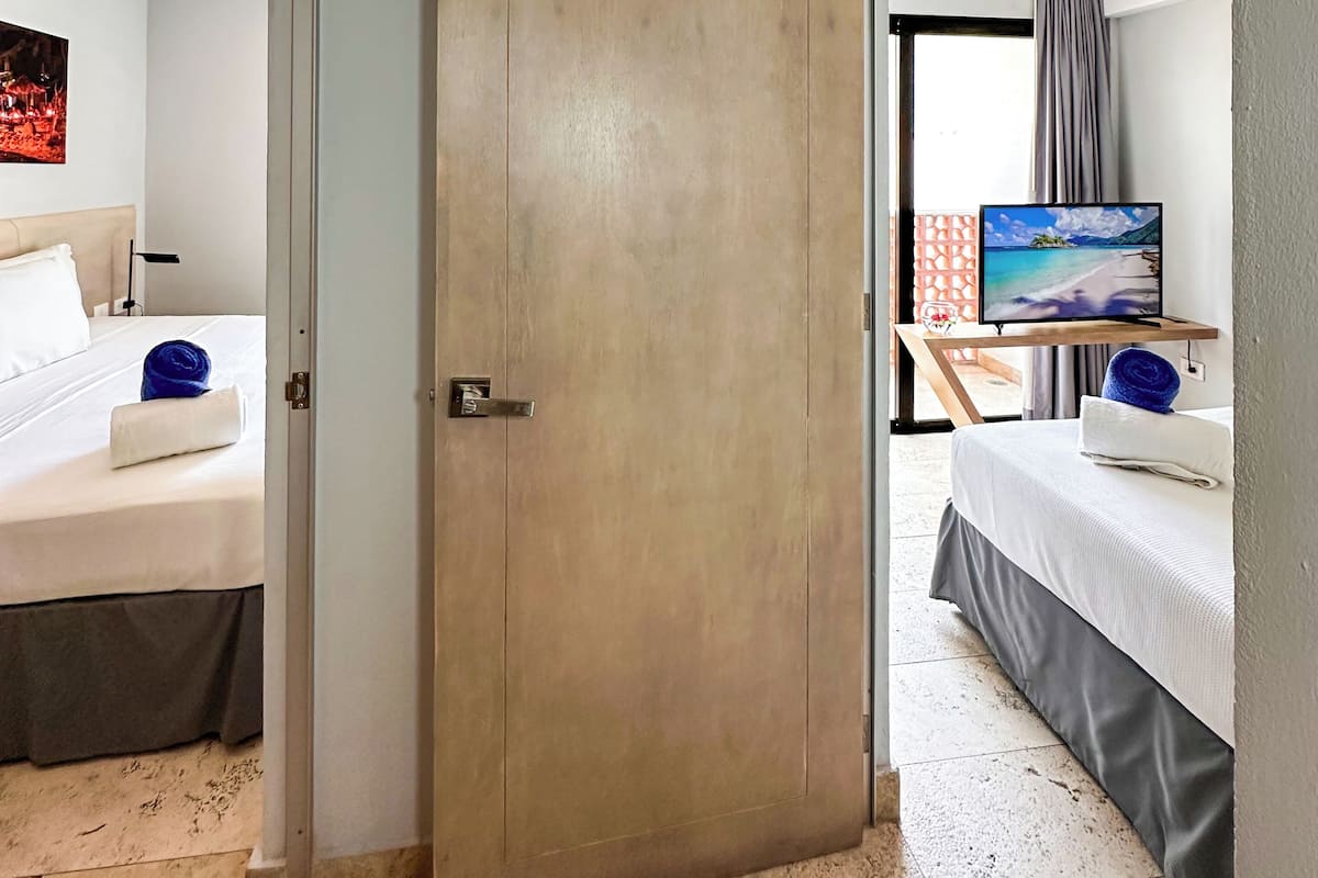 A hotel room with a large wooden door, a bed with a white comforter, a flat-screen TV on a wooden stand, and a view of a beach through the window.