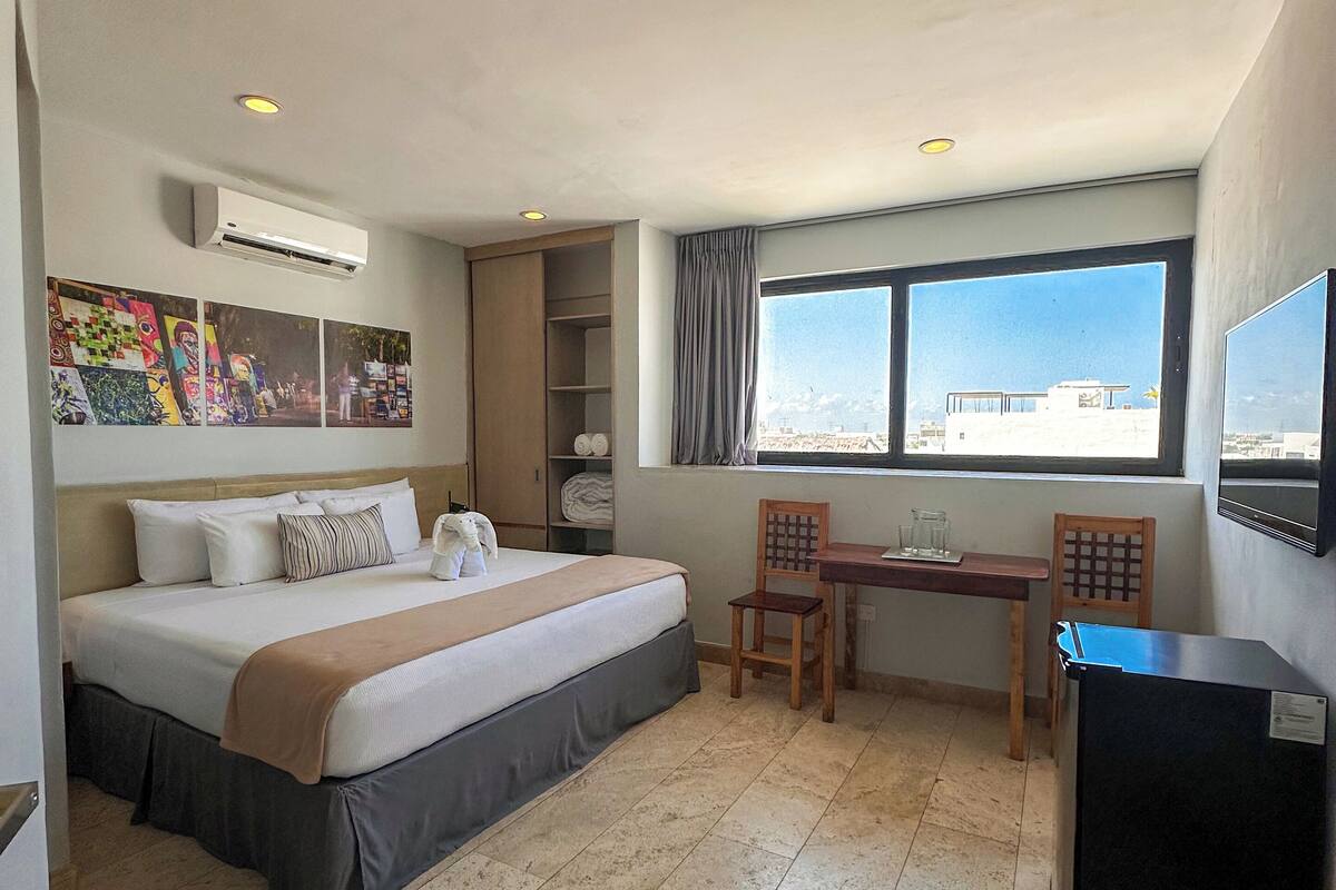 A hotel room with a bed, a desk, a chair, a TV, and a window with a city view.
