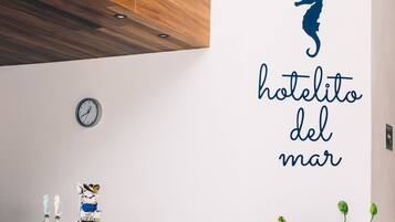 Un interior de hotel moderno con techo de madera, pared blanca con un caballito de mar y texto, escritorio con sillas, un reloj y una planta en maceta.