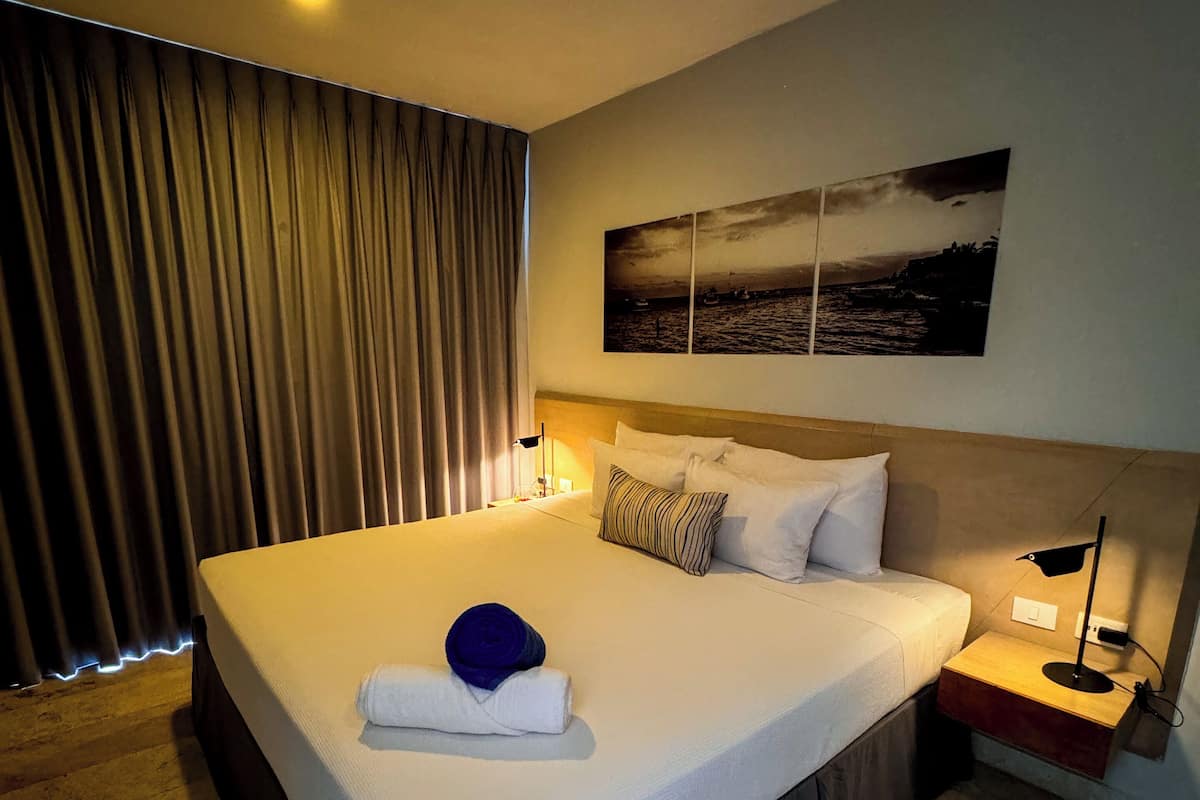 A hotel room with a large bed, a nightstand with a lamp, a painting of a seascape, and a view of the sea through the window.