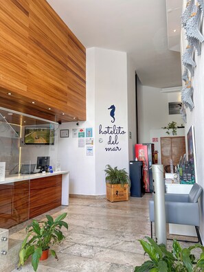 Un hall d’hôtel avec un plafond en bois, un comptoir d’accueil, un mur orné du logo d’un hippocampe et de l’inscription « hotelito del mar », un réfrigérateur Coca-Cola et des plantes en pot.