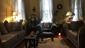 A cozy living room with two sofas, a coffee table, and a floor lamp. There is a large window with curtains, a wall clock, and a potted plant. - Historic Iron Horse Inn (Hermann)