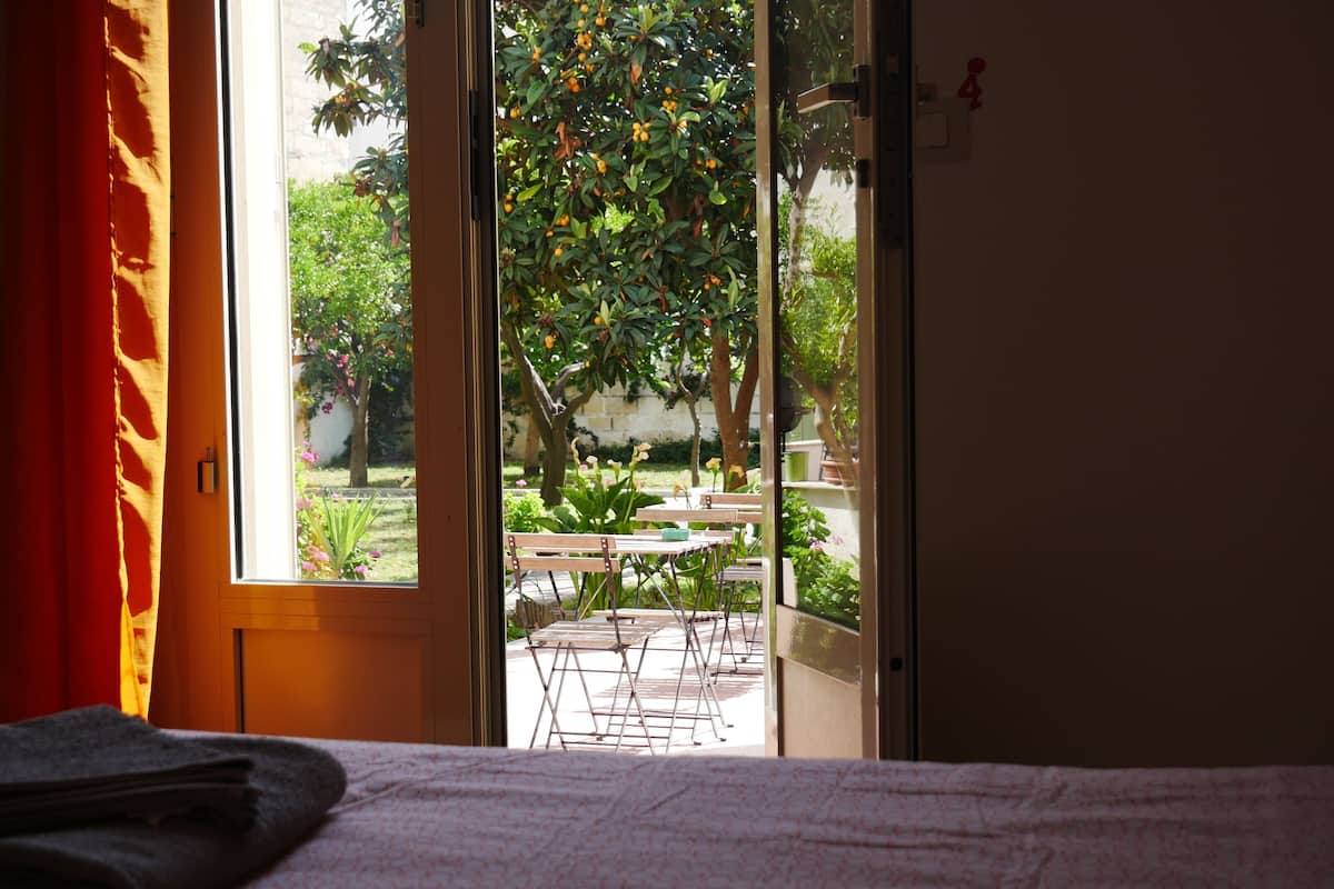 Una habitación con cama, ventana y vista a un jardín exterior con mesas y sillas.