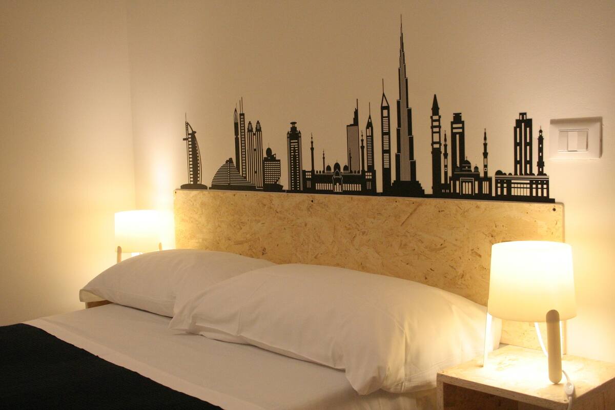 Una habitación de hotel con una cama, dos lámparas de noche y una silueta decorativa del horizonte en la pared.