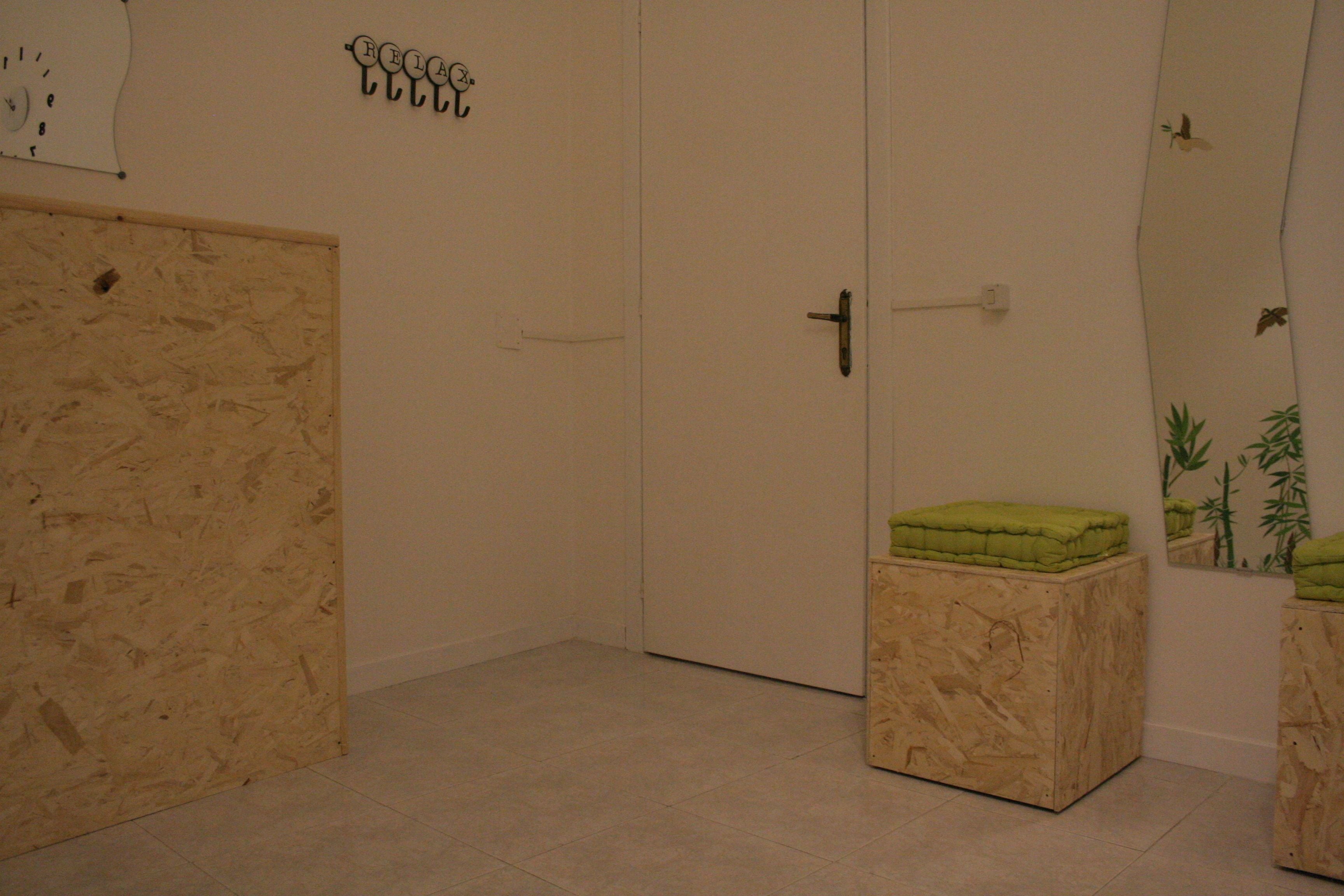 A room with a wooden door, a white wall with a clock, and a green cushion on a wooden stool.