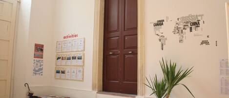 A hallway with a brown door, a bulletin board with various notices, a potted plant, and a wooden chest.