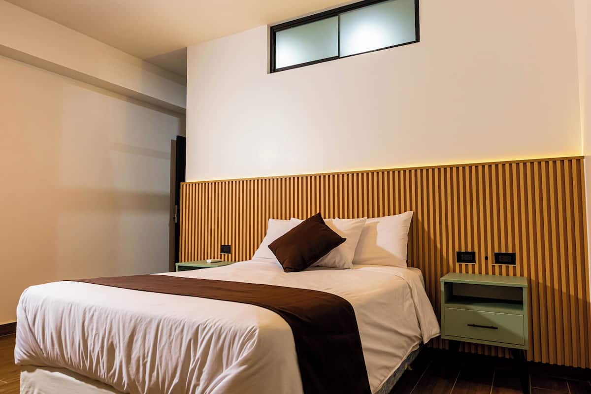 A neatly made bed with a brown accent, a bedside table with a lamp, and a wall-mounted light fixture.