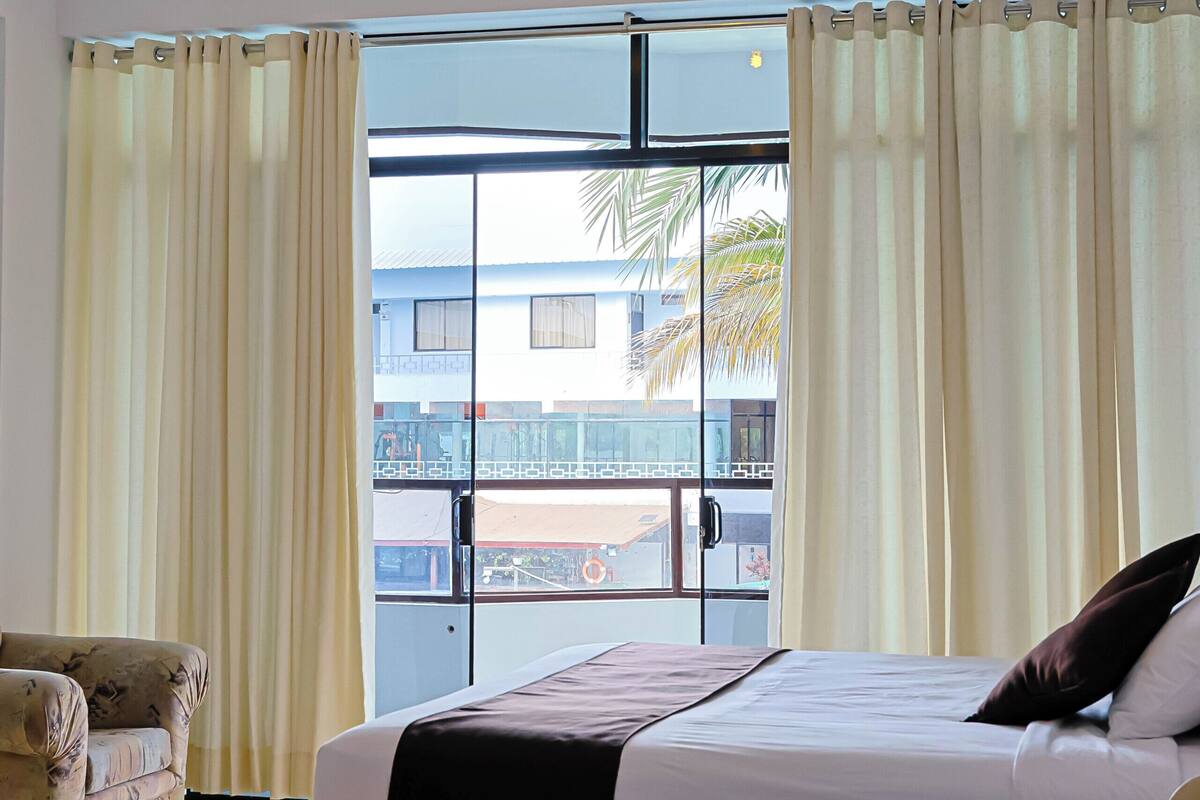 A hotel room with two beds, a sofa, a window with curtains, and a view of a building and palm trees outside.