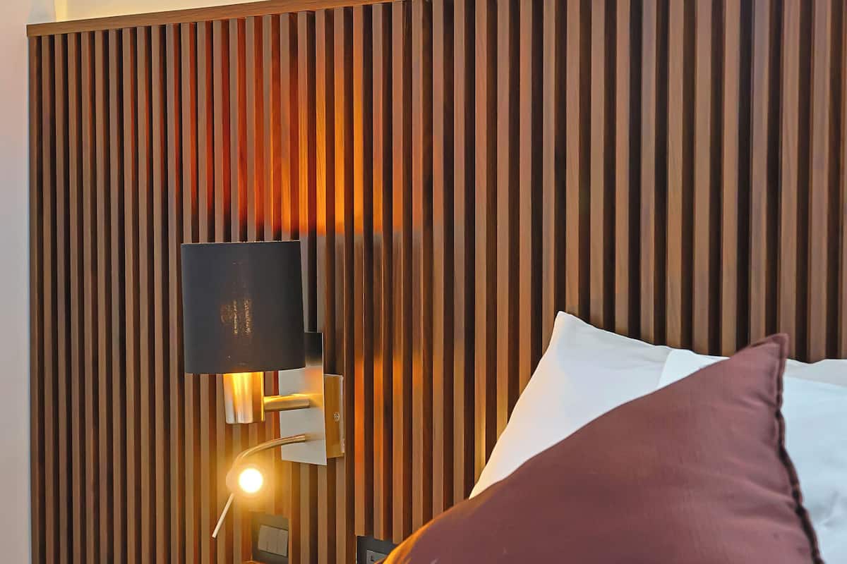 A neatly made bed with white and brown pillows, a wooden slatted headboard, and a wall-mounted light fixture.