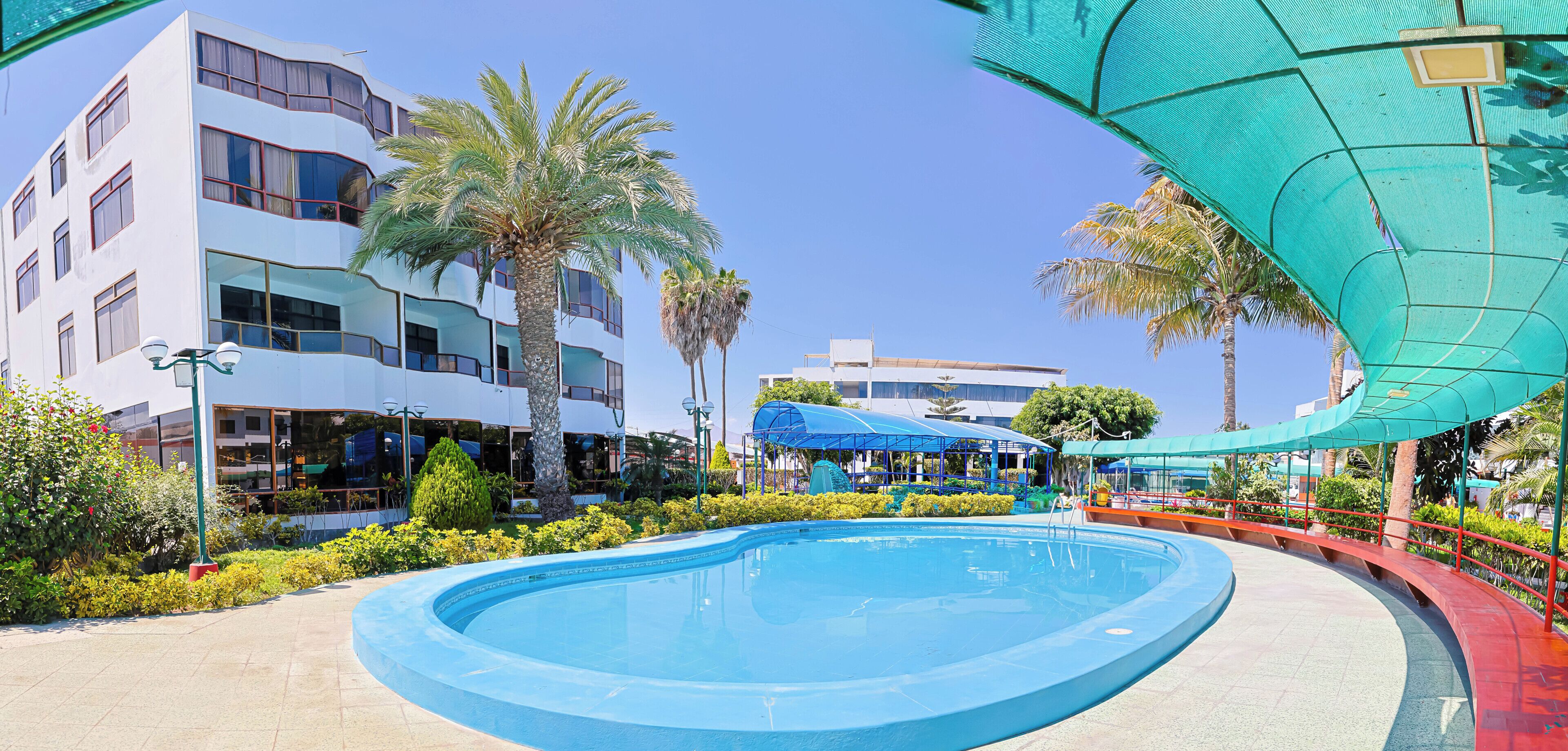 A swimming pool with a curved edge, surrounded by greenery and a building with multiple floors and balconies.