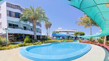 A swimming pool with a curved edge, surrounded by greenery and a building with multiple floors and balconies.