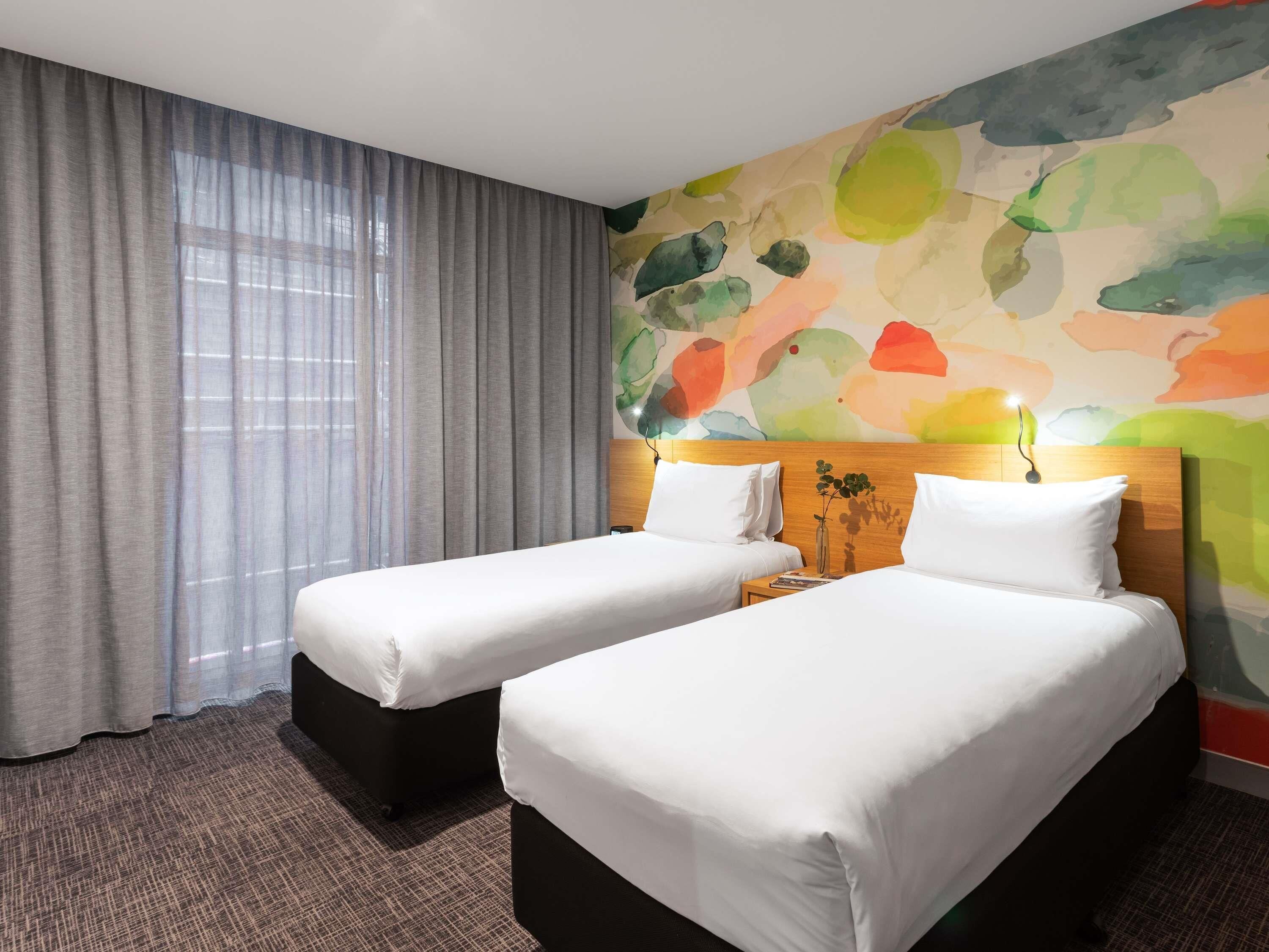 A hotel room with two beds, a large window with sheer curtains, and a colorful abstract wall mural.
