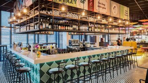 Una zona de bar con mostrador, taburetes, estantes con botellas e iluminación colgante.