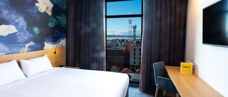 A hotel room with a large bed, a desk, and a view of a cityscape through a large window.