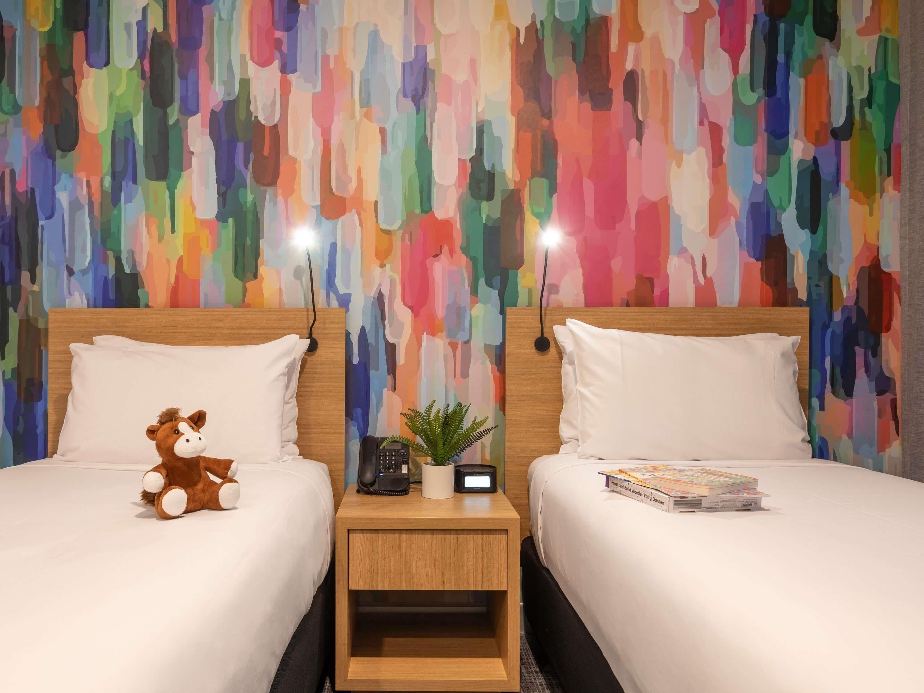 Habitación de hotel con dos camas, cada una con almohadas blancas y cabeceros de madera. Un peluche está en la cama de la izquierda, y una pequeña planta y un libro en la de la derecha. La pared detrás de las camas presenta un diseño abstracto y colorido.