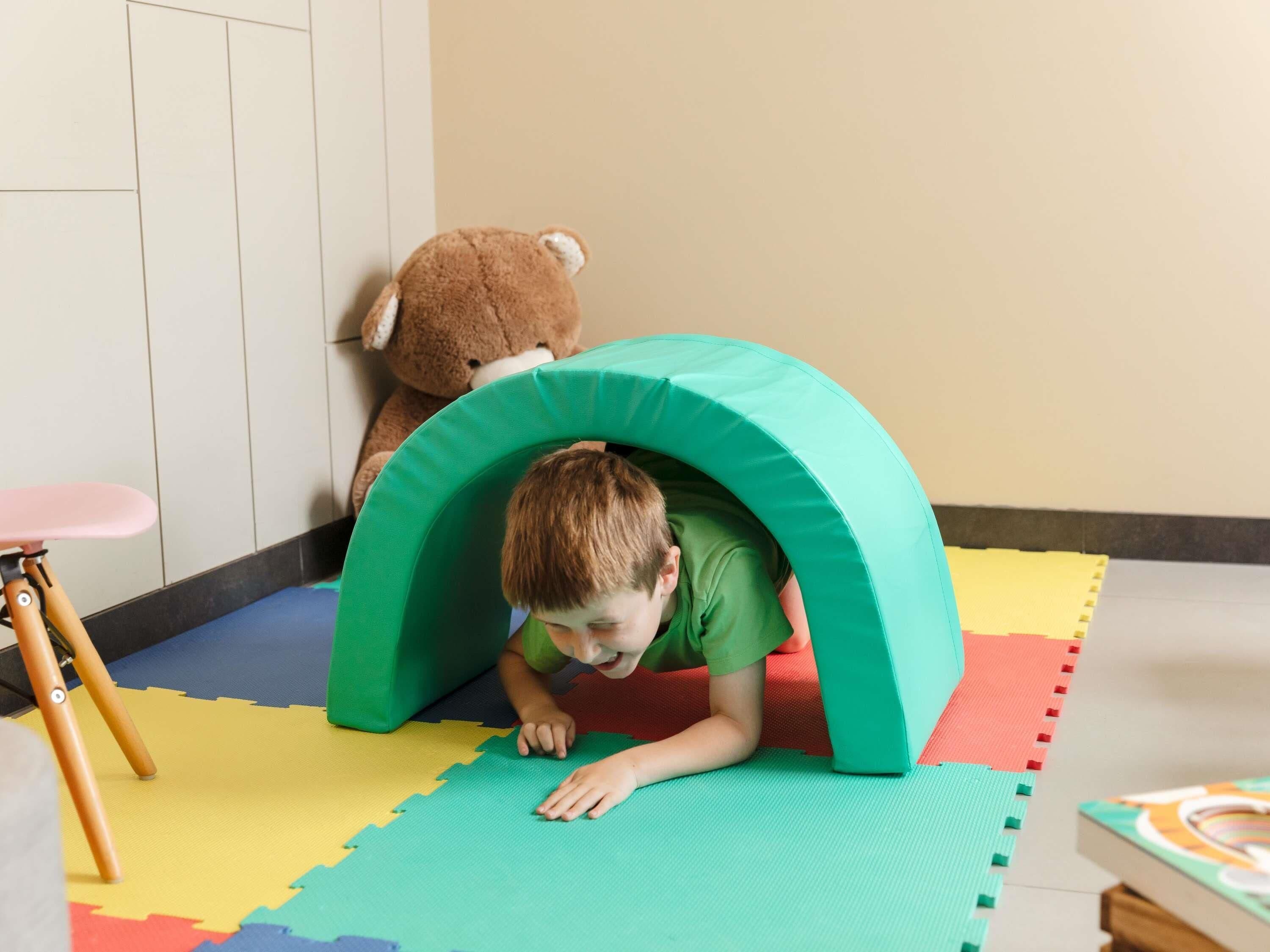 Un niño jugando debajo de un túnel verde en una habitación con un tapete de juegos de colores, un osito de peluche y una silla rosa.