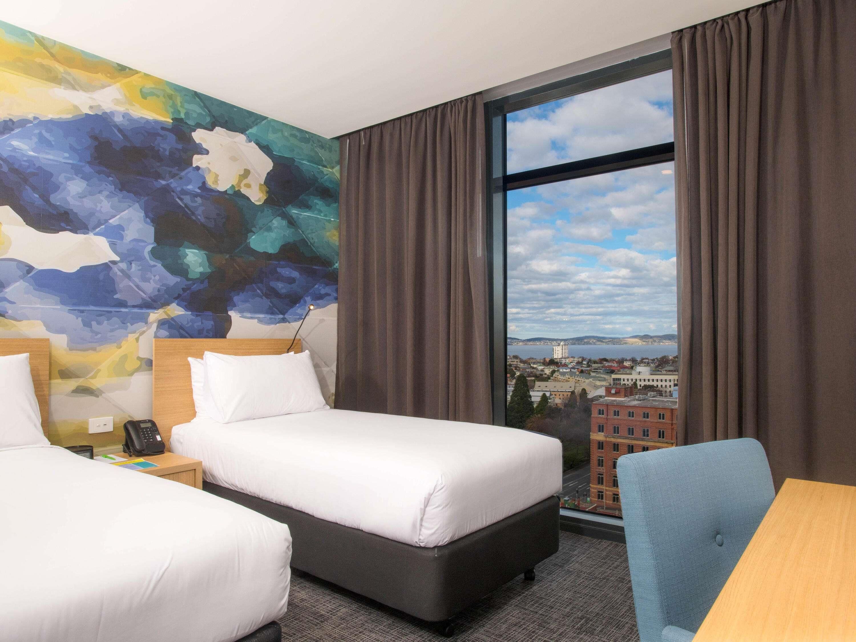 A hotel room with two beds, a large window with a city view, a colorful abstract wall, and a small table.