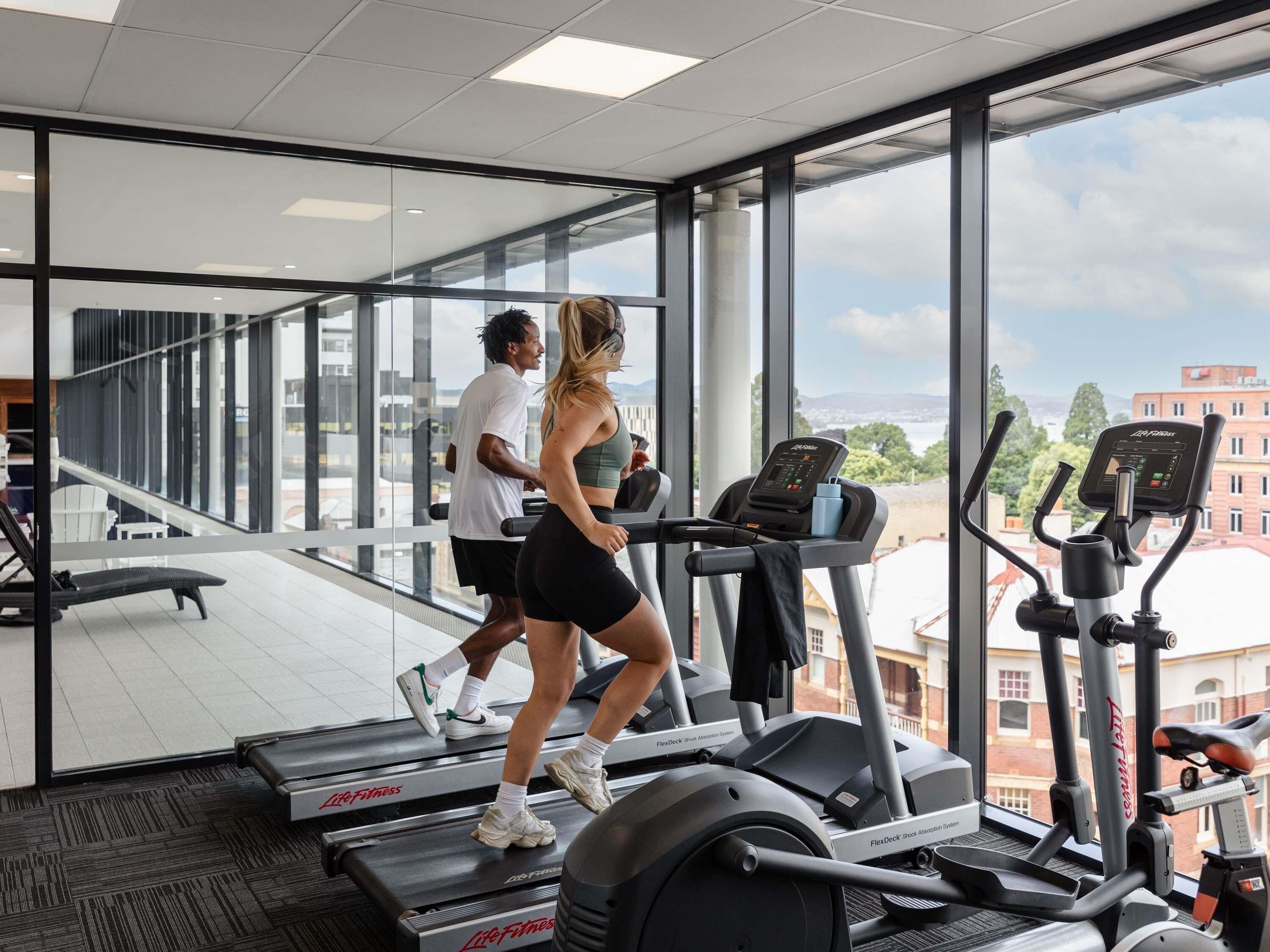 A gym with treadmills and elliptical machines, large windows, and a view of buildings and sky.