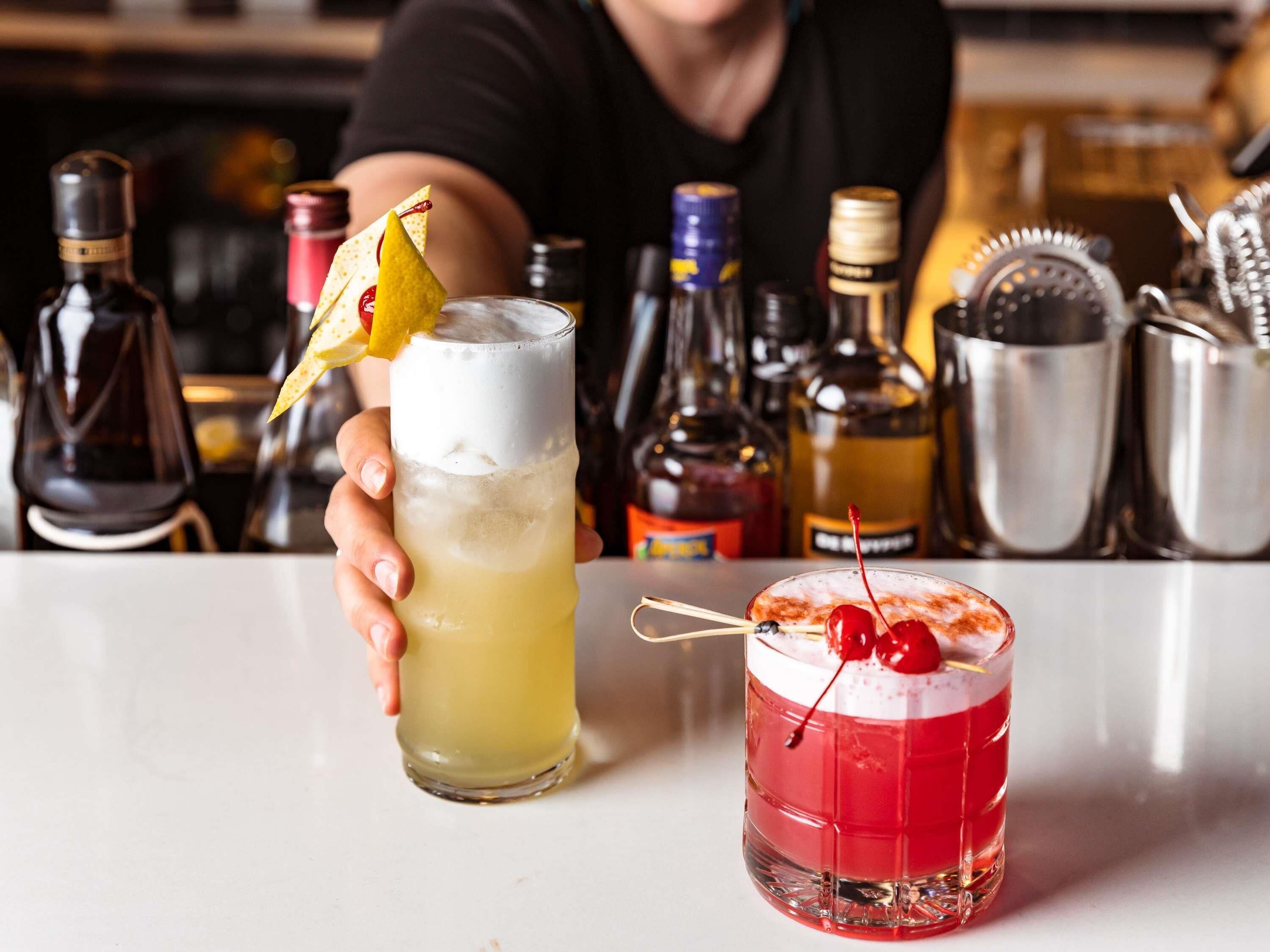 Un barman sosteniendo dos bebidas, una en un vaso alto y otra en uno bajo, con varias botellas y un shaker al fondo.