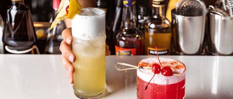 A bartender holding two drinks, one in a tall glass and one in a short glass, with various bottles and a shaker in the background.