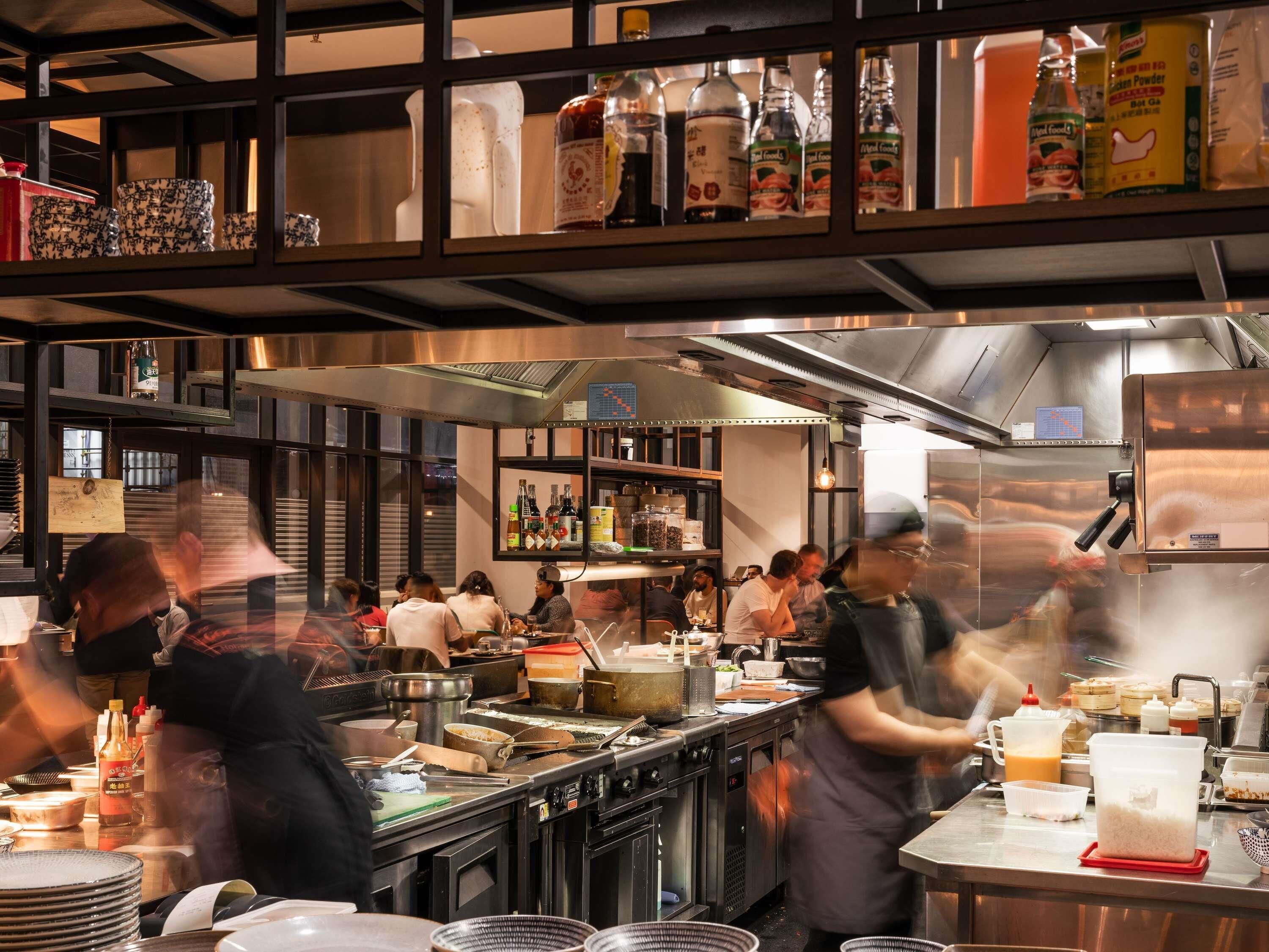 A busy kitchen with chefs preparing food, stainless steel appliances, and shelves stocked with condiments.