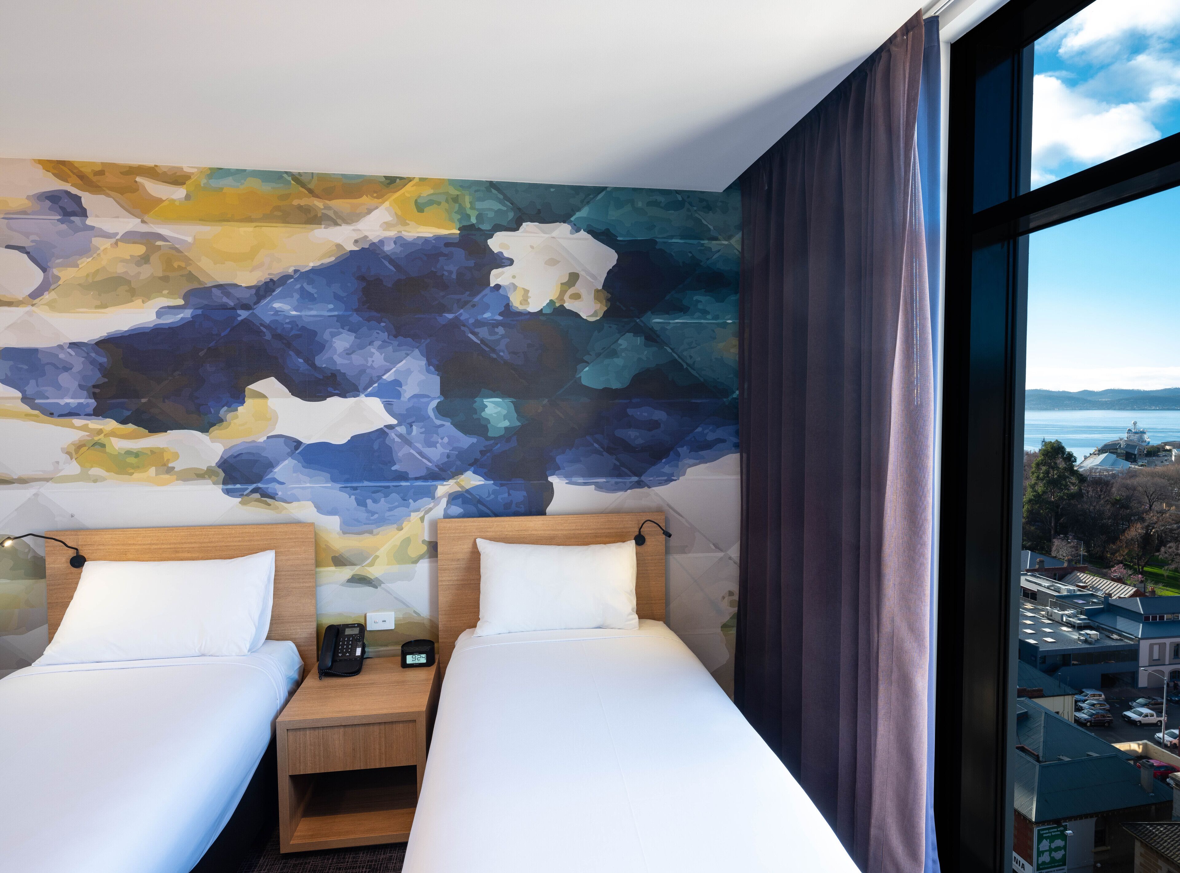 A hotel room with two beds, a large abstract mural on the wall, a view of a cityscape through a window, and a wooden headboard.