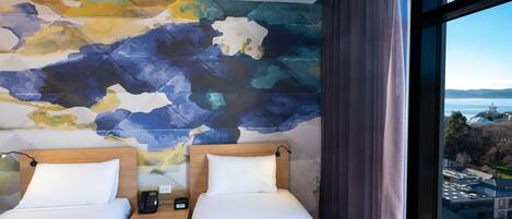 Habitación de hotel con dos camas, un gran mural abstracto en la pared, vistas de un paisaje urbano a través de una ventana y un cabecero de madera.