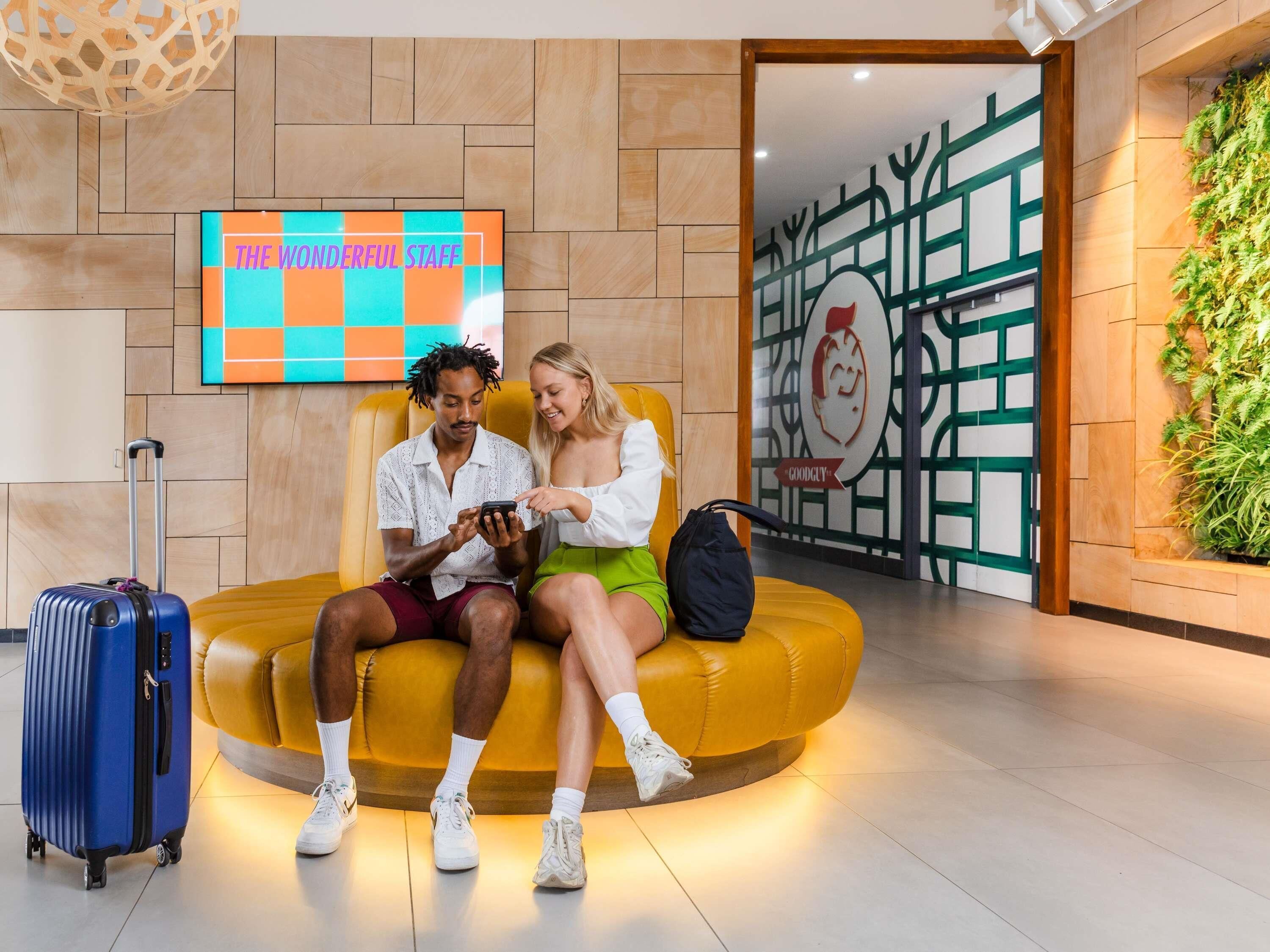 A couple sitting on a yellow sofa in a lobby with a 'The Wonderful Staff' sign, a wall-mounted TV, and a green wall with a logo.