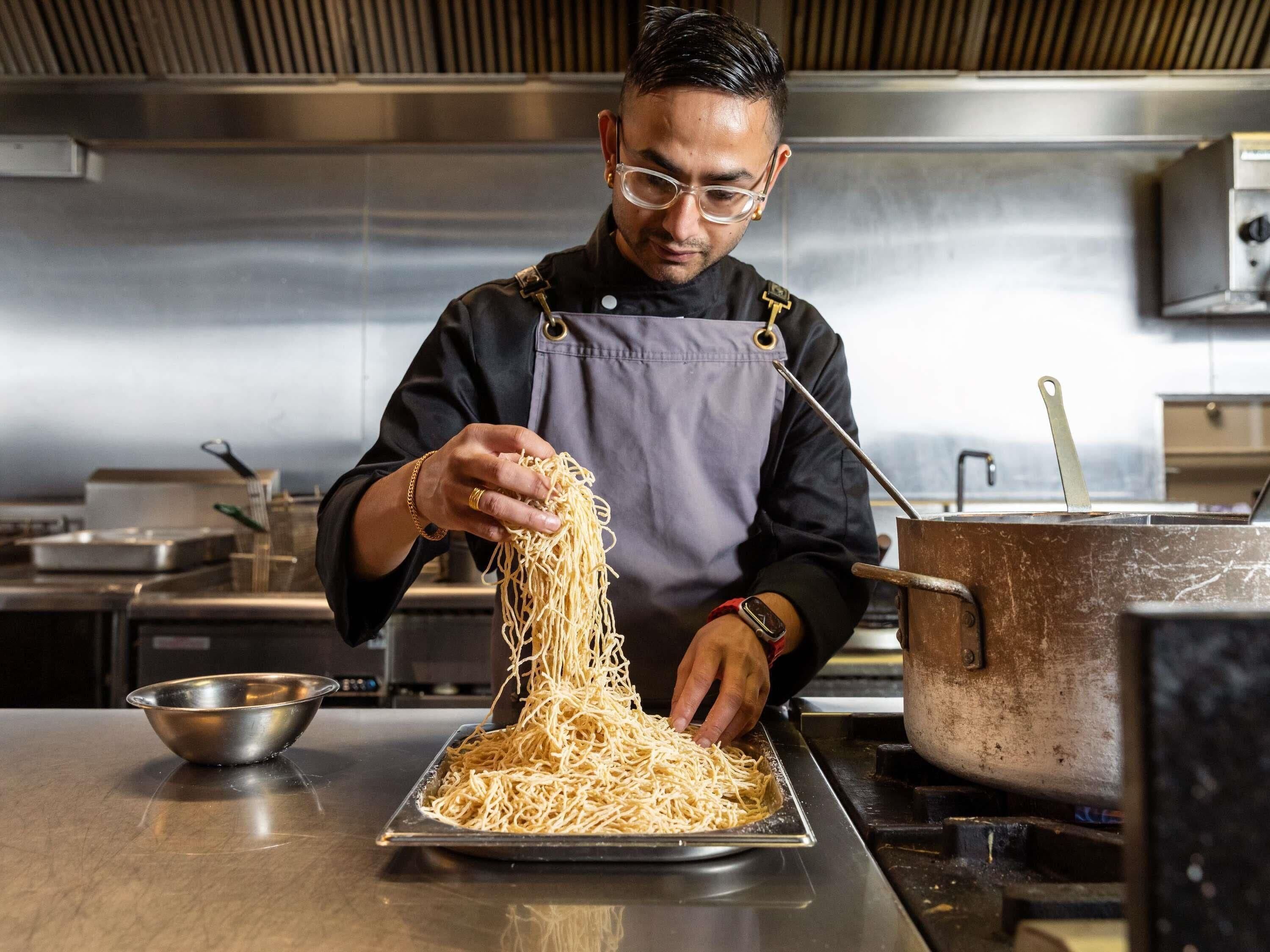 A chef preparing noodles in a professional kitchen with stainless steel surfaces and modern appliances.