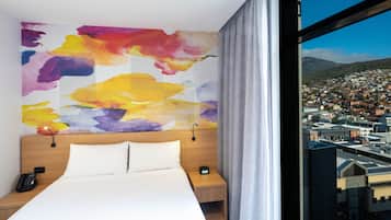 Une chambre d’hôtel avec un grand lit, une œuvre d’art abstraite aux couleurs vives accrochée au mur, une vue sur le paysage urbain par une grande fenêtre, et une table de chevet avec un téléphone et une horloge.