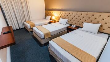 A hotel room with two single beds and one double bed, a bedside table with a lamp, a phone, and a remote control on a shelf.