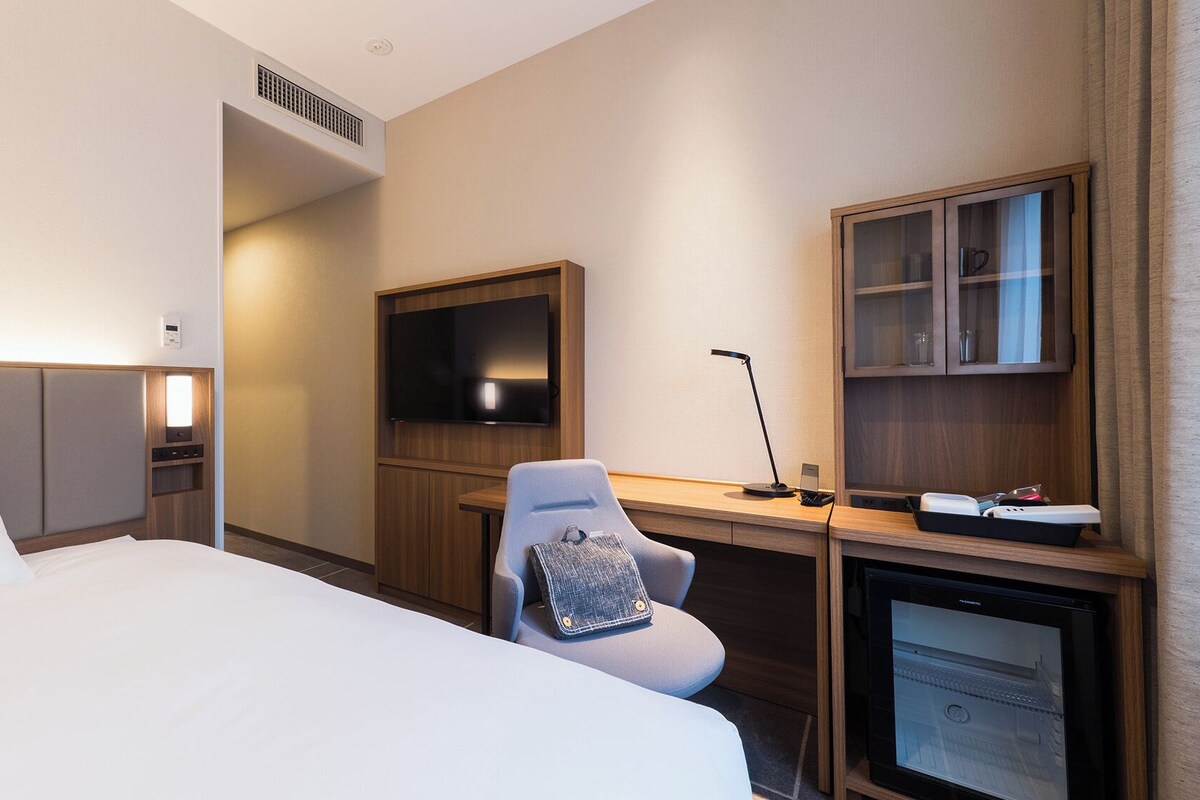 A hotel room with a bed, a desk with a TV, a chair, a desk lamp, a minibar, and a wall-mounted TV.