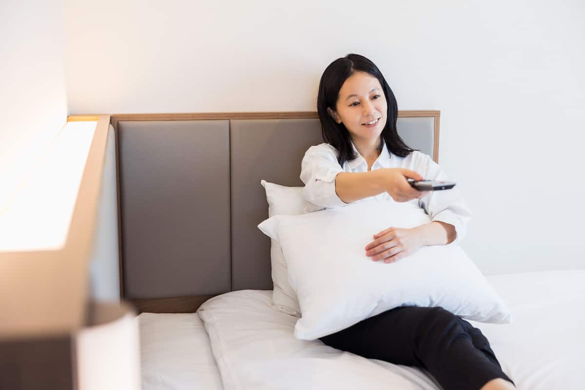A person sitting on a bed with a white pillow, holding a remote control.