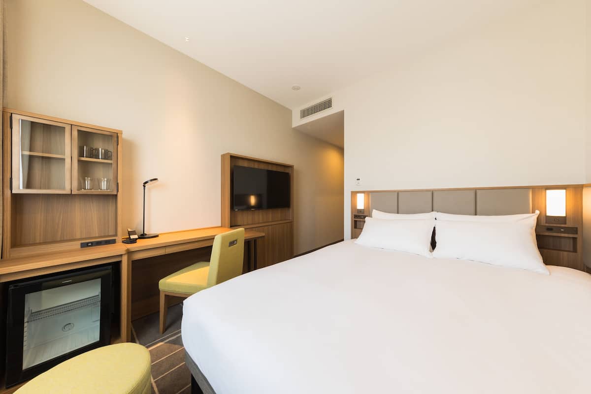 A hotel room with a bed, a desk with a chair, a television, and a minibar.
